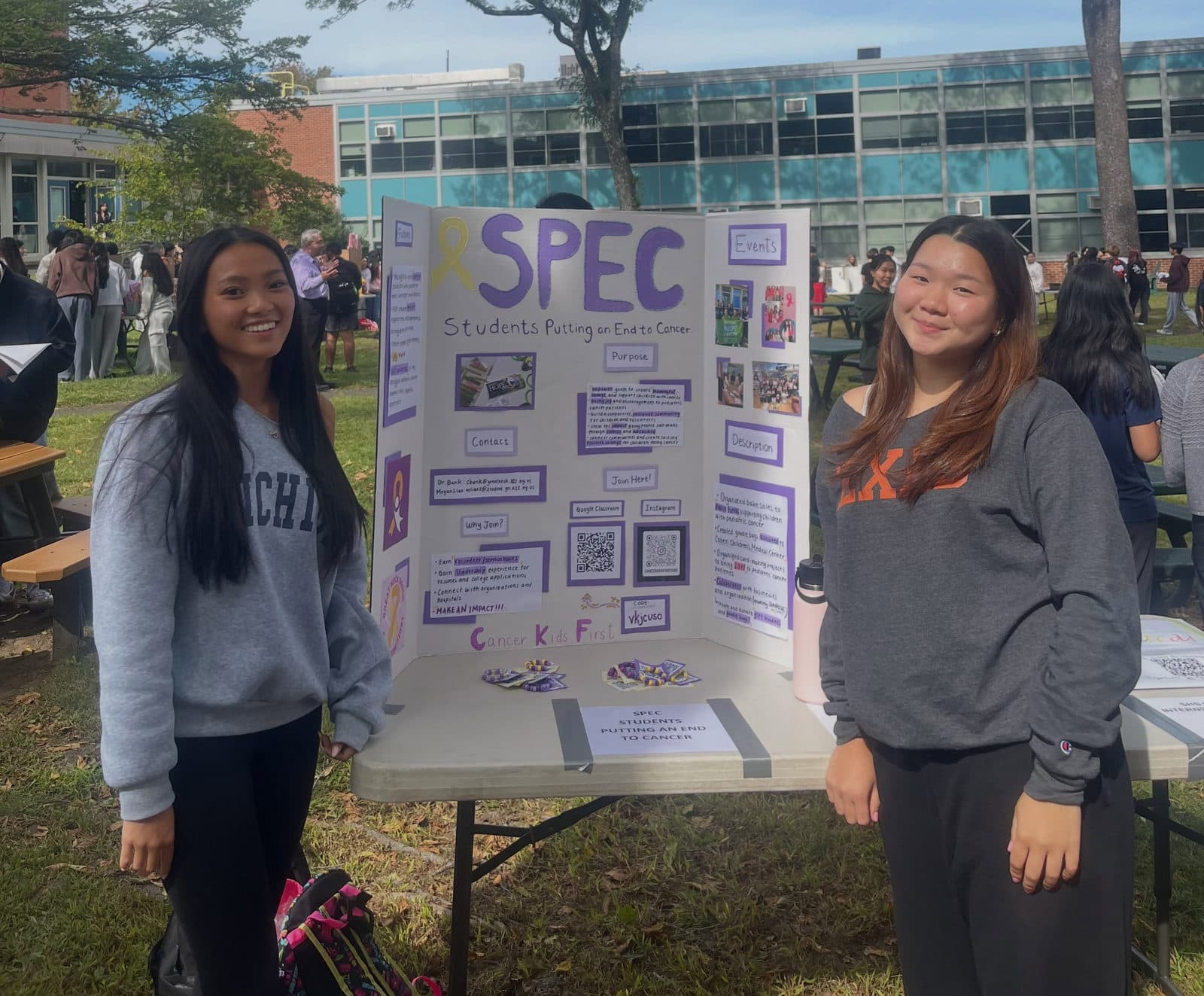 Two students standing on either side of a hand-made tri-fold display board headed 'SPEC - Students Putting an End to Cancer', set up on a folding table on grass, with other stalls and a school building behind them.