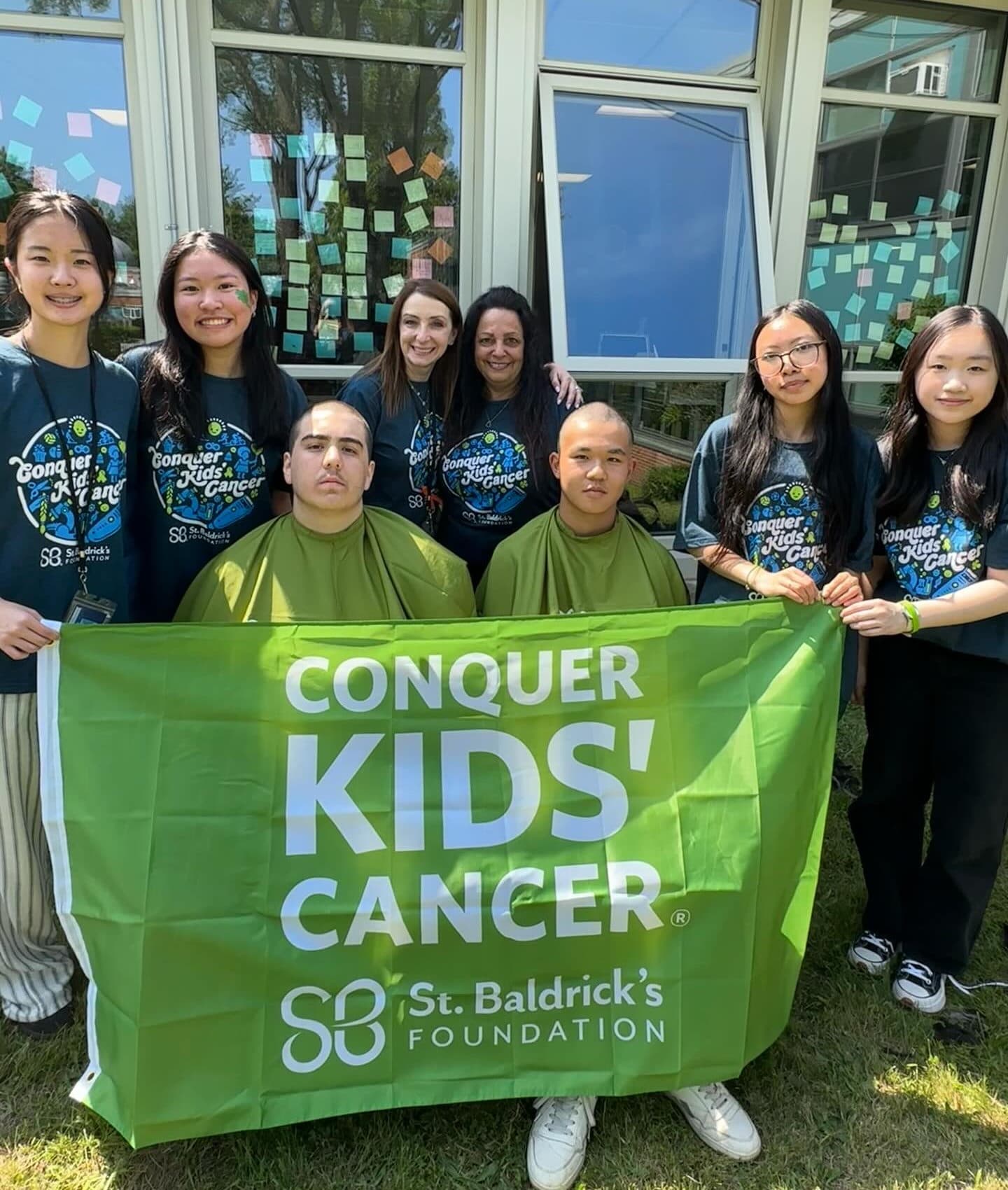 Two people with freshly shaved heads sit in green barber capes, holding up a large green banner lettered Conquer Kids' Cancer above the St. Baldrick's Foundation name and mark. Others in matching printed T-shirts stand behind them on the grass, outside a school building whose windows are covered in small coloured paper notes.