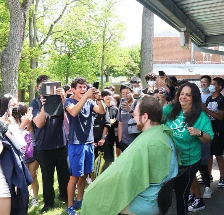 Outdoors under a walkway canopy, a seated person in a green barber cape has their head shaved by someone in a green St. Baldrick's Foundation T-shirt, while a crowd of students, several of them in face masks, watches and films on phones.