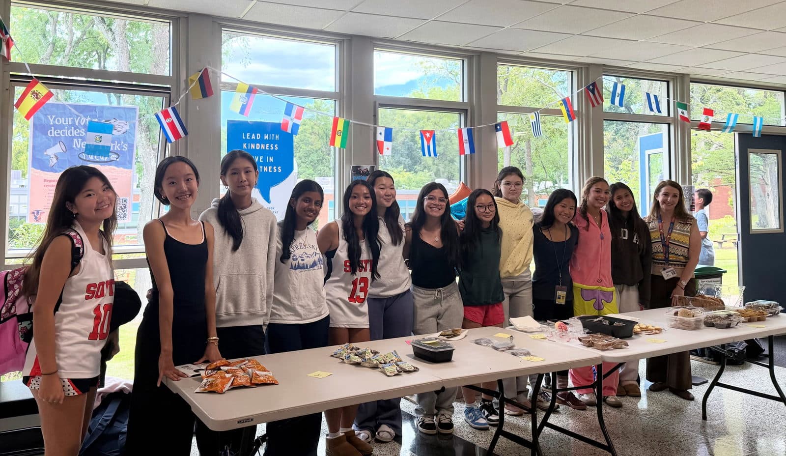 A long row of students standing shoulder to shoulder behind a folding table laid with wrapped baked goods, in a school lobby hung with a line of small national flags and lit by tall windows.