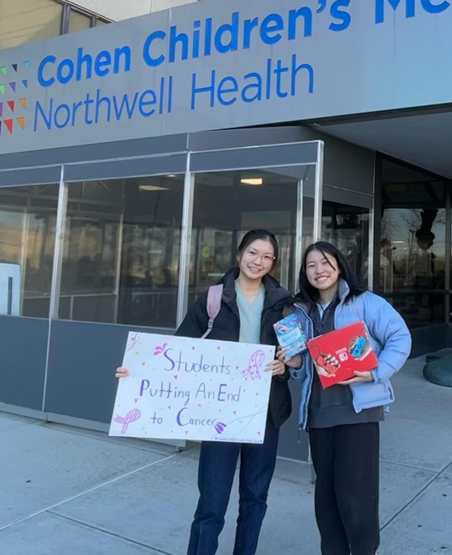 Two students standing on the pavement outside an entrance signed Cohen Children's Medical Center, Northwell Health. One holds a hand-lettered poster reading 'Students Putting An End to Cancer'; the other carries two wrapped gift boxes.